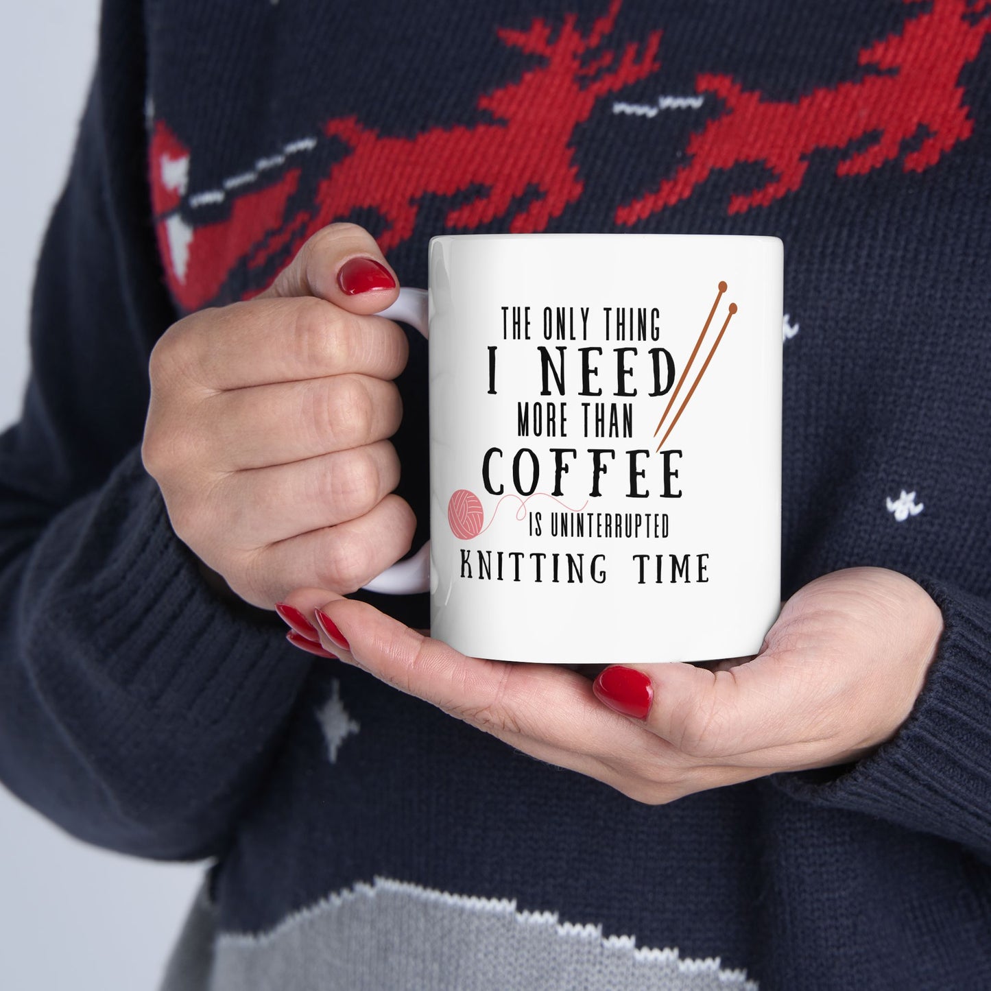 Person holding a mug with a knitting-themed quote "The Only Thing I Need More Than Coffee Is Uninterrupted Knitting Time", wearing a black sweater with red deer pattern.
