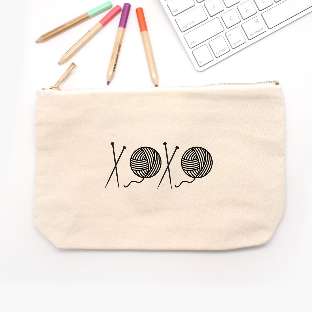 Beige pouch with yarn and knitting needle design on a white surface with markers and a keyboard.