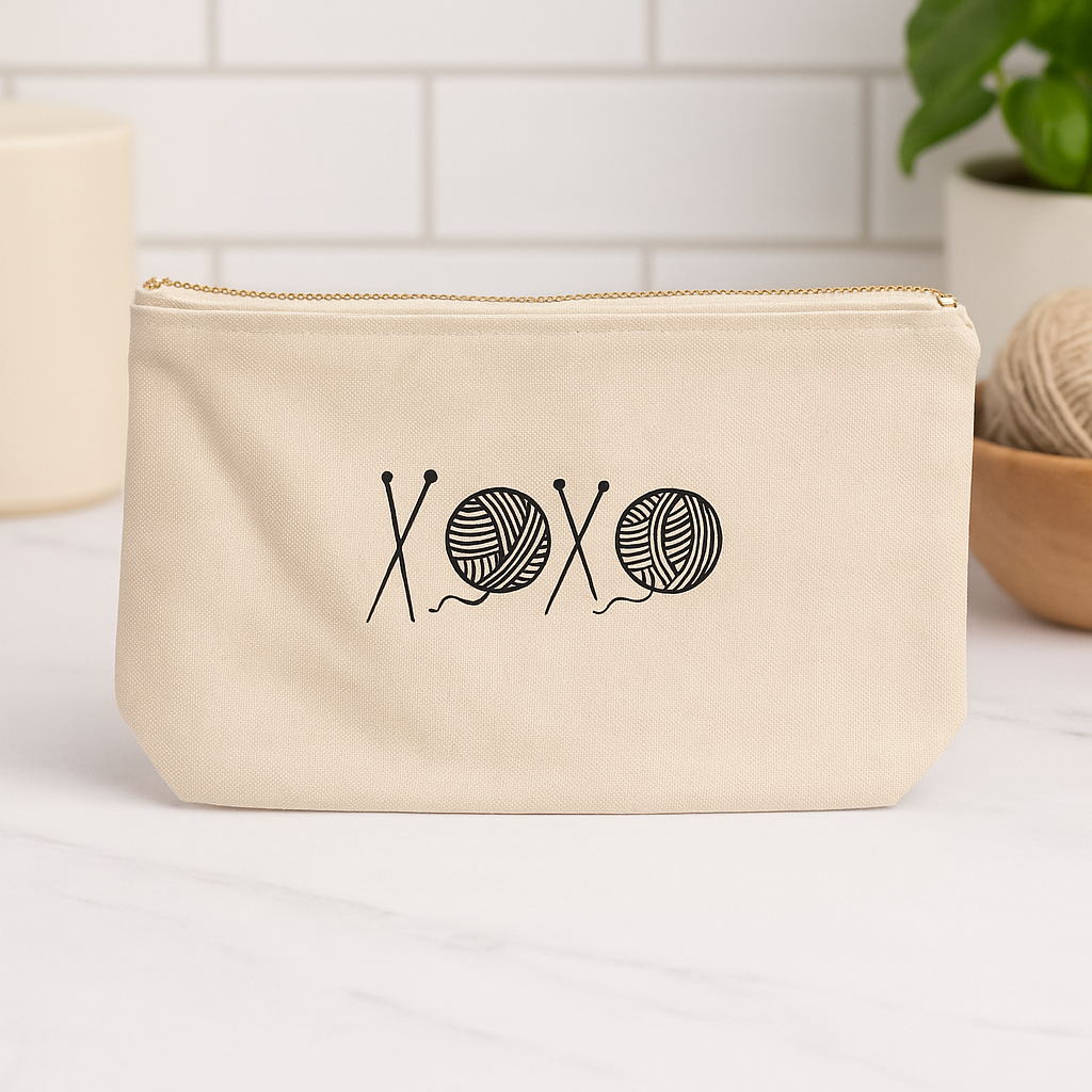 Beige zippered canvas notions bag featuring a black "XOXO" design with knitting needles and yarn balls, displayed on a white countertop with yarn and a potted plant in the background.