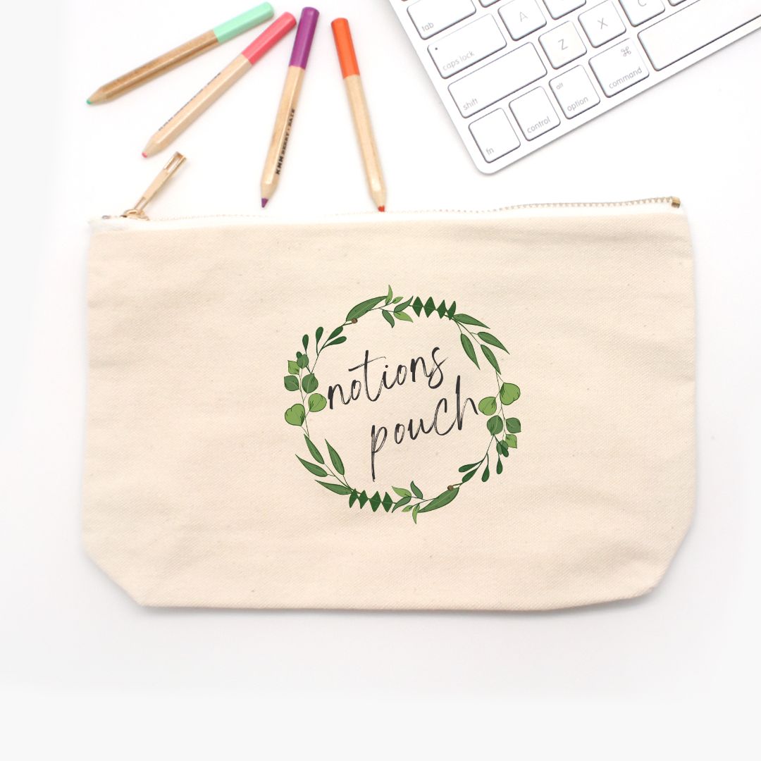 A beige canvas notions bag on a white table near a white keyboard and markers. The bag has a design on the front with green leaves in a circle and says "Notions Pouch" in the center. A great notions bag for knitters and crocheter