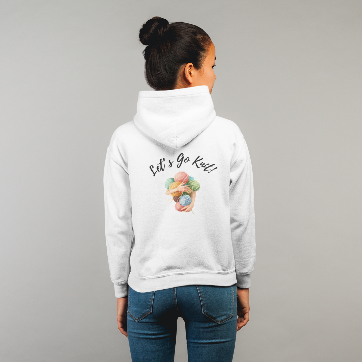 A woman wearing blue jeans and a white hoodies that says "Lets Go Knit" on the back. The design also has a pair of hands holding a lot of yarn. She is standing in front of a white wall.
