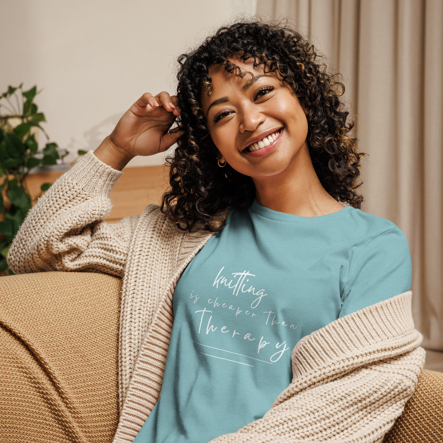 Woman wearing a white sweater over a blue t-shirt with text, sitting on a couch. The shirt says "Knitting Is Cheaper Than Therapy".