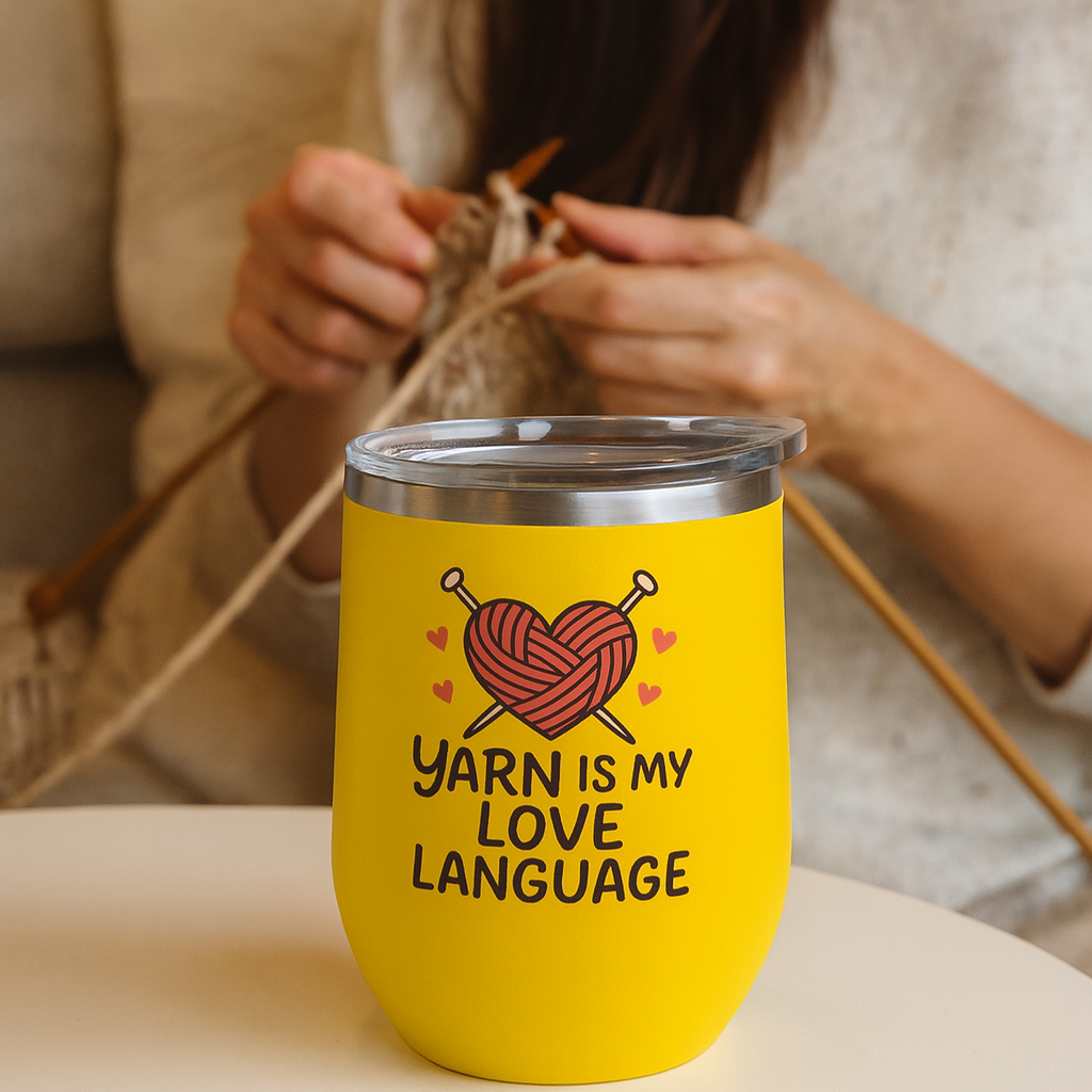 Yellow tumbler with 'Yarn is my love language' text and heart design on a table with a person knitting in the background.