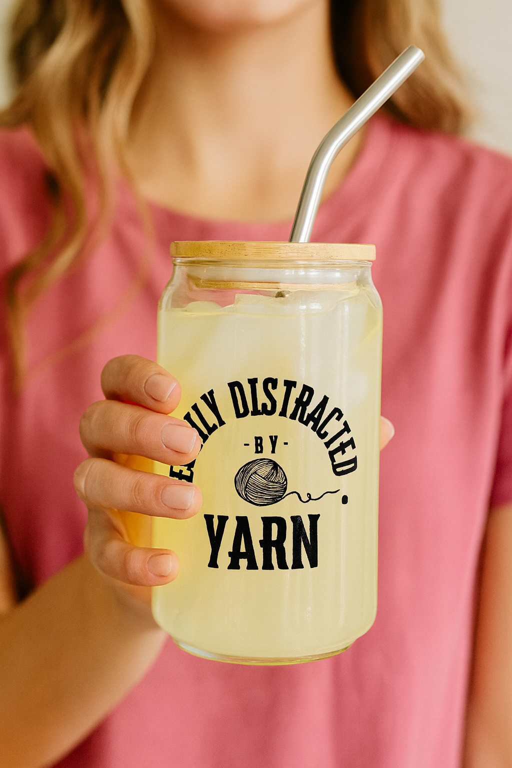 Person holding a glass jar with a straw, labeled 'Easily Distracted by Yarn', against a pink shirt background.