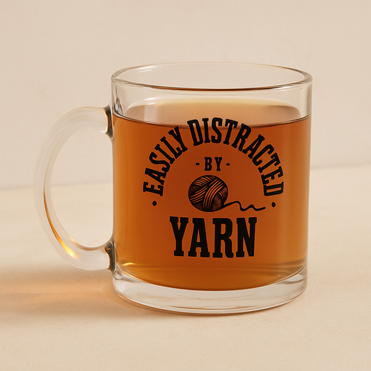 Clear mug with 'Easily Distracted by Yarn' text and yarn ball graphic filled with a warm beverage on a beige background.