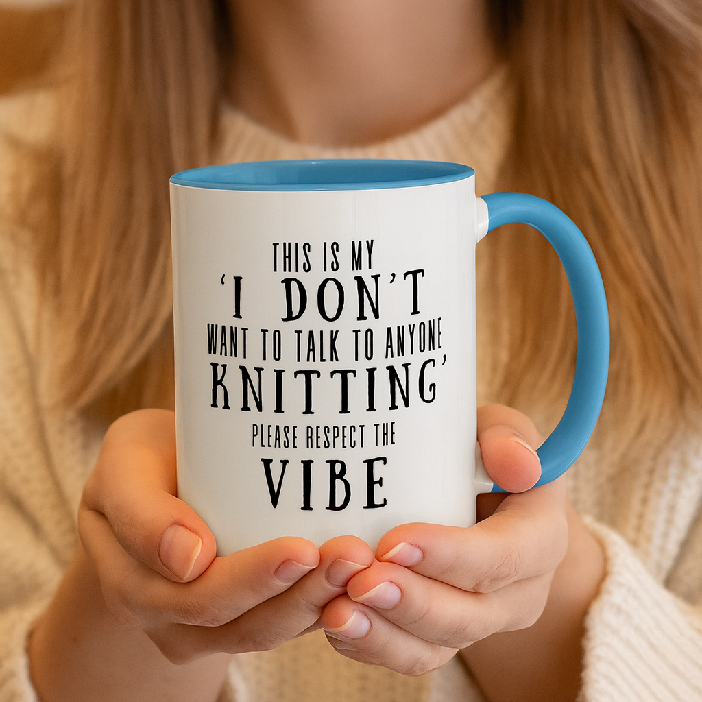 Person holding a  white mug with text in black letters that says This Is My I Don't Want To Talk To Anyone Knitting Please Respect The Vibe.  The rim and handle of the mug is light blue.