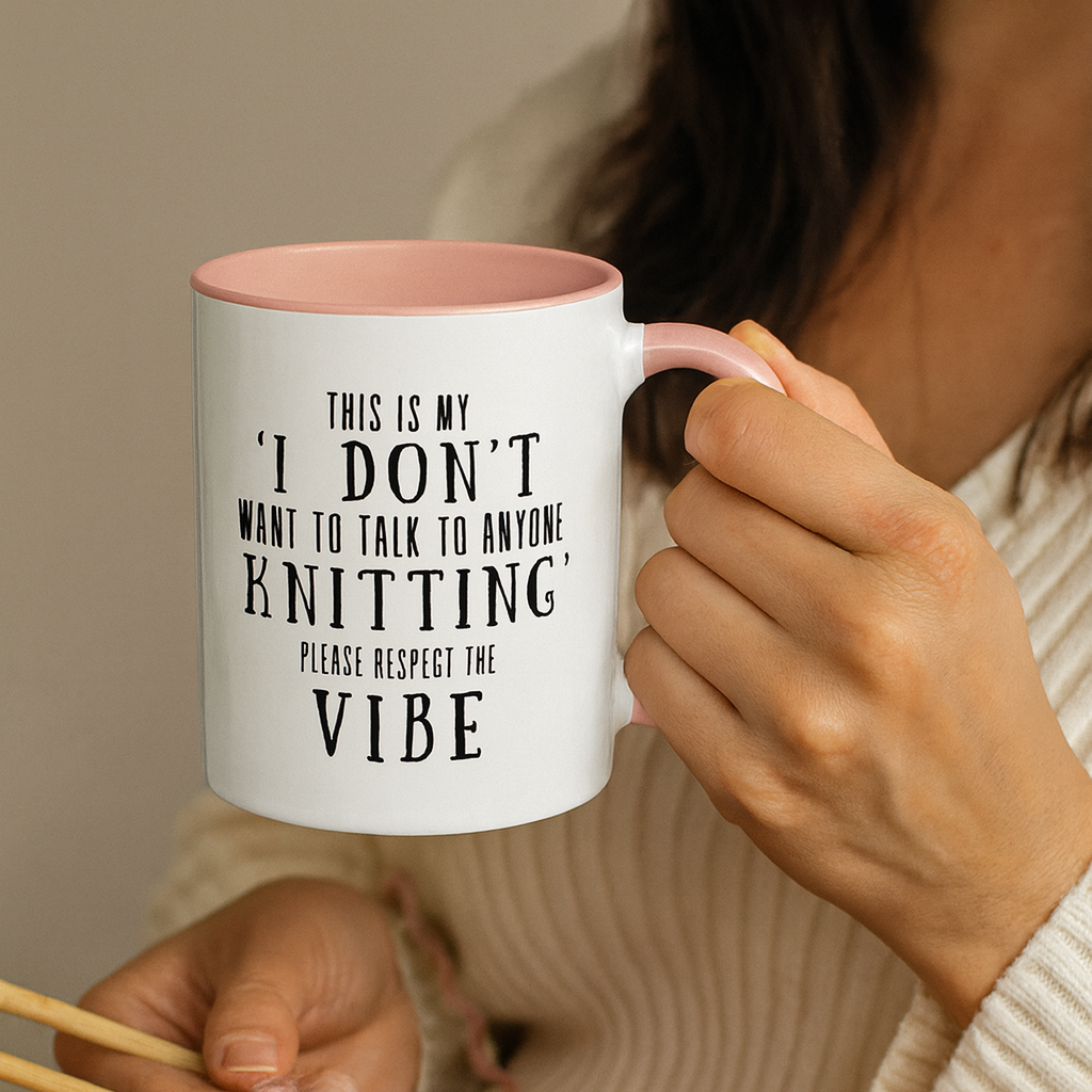 A person holding a white mug with text that says "This Is My I Don't Want To Talk To Anyone Knitting Please Respect The Vibe".  The text is in black font, and the rim and handle is light pink in color.