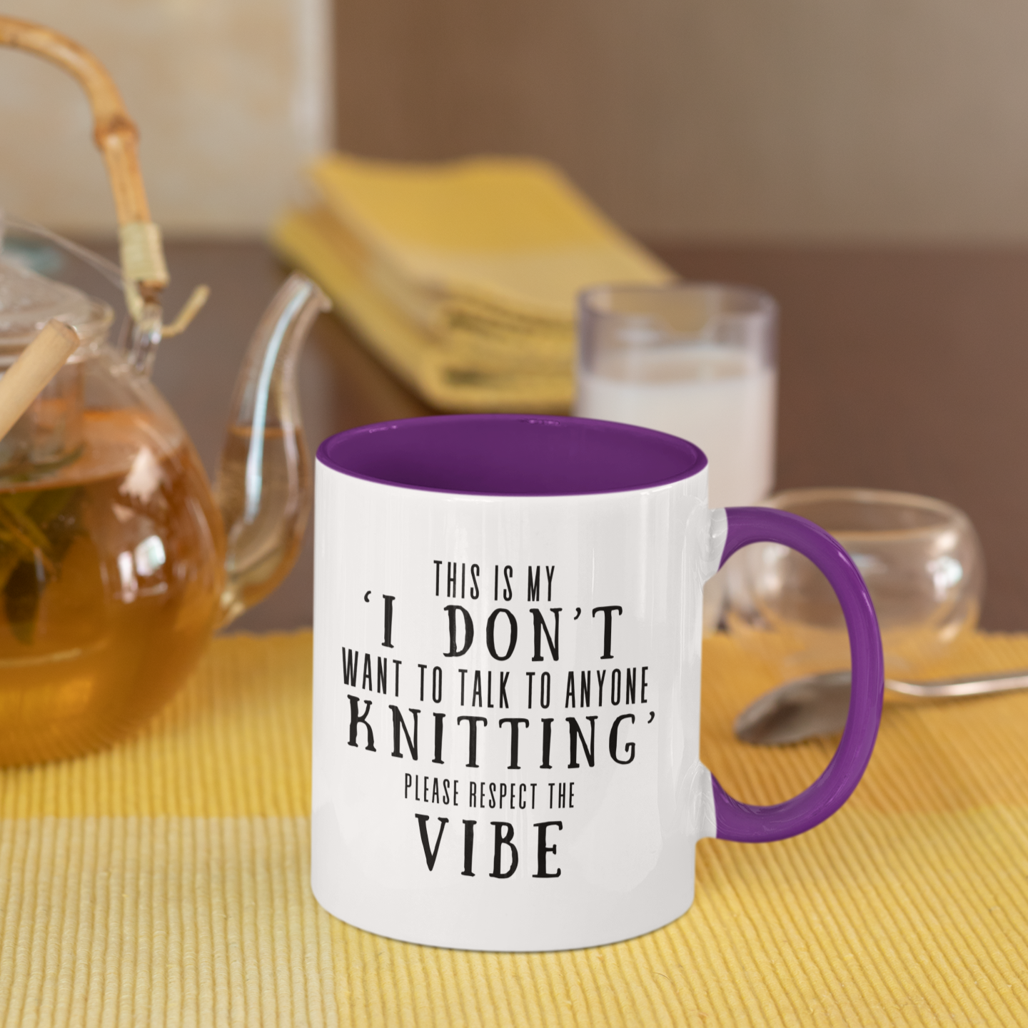 A white mug with purple interior and handle on a table with tea and snacks.  The mug says "This is my  I Don't Want To Talk To Anyone Knitting Please Respect The Vibe" in black letters.