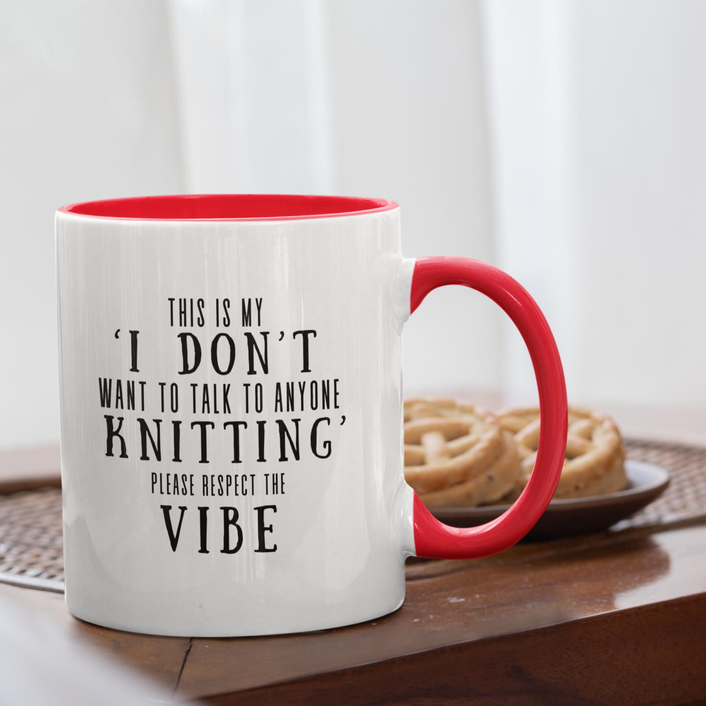 A white mug with red rim and handle, that says "This is my I don't  want to talk to anyone knitting please respect the vibe", in black letters.  The mug is on a brown wooden table, with a plate of cookies behind it.