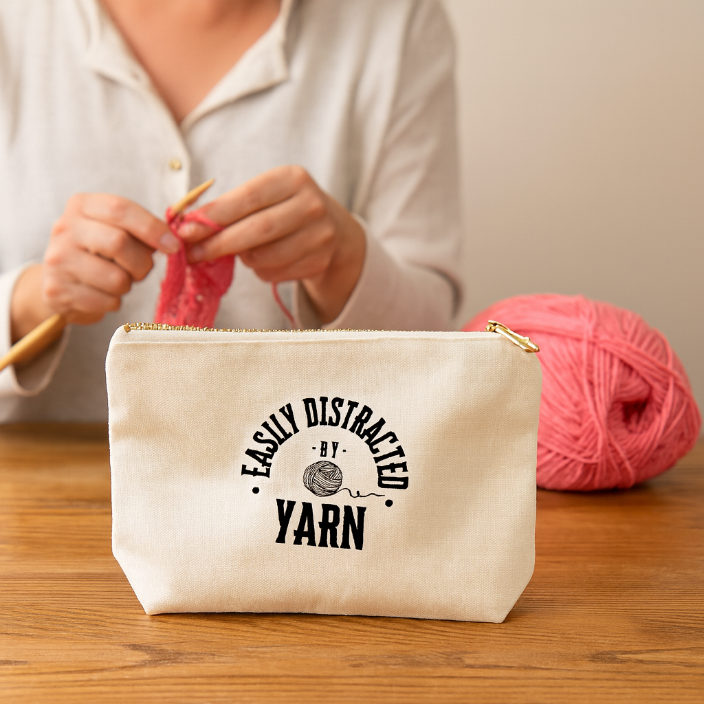 Person knitting with a pink yarn and a natural color notions bag that says  'Easily Distracted by Yarn'.  The bag is on a wooden table.
