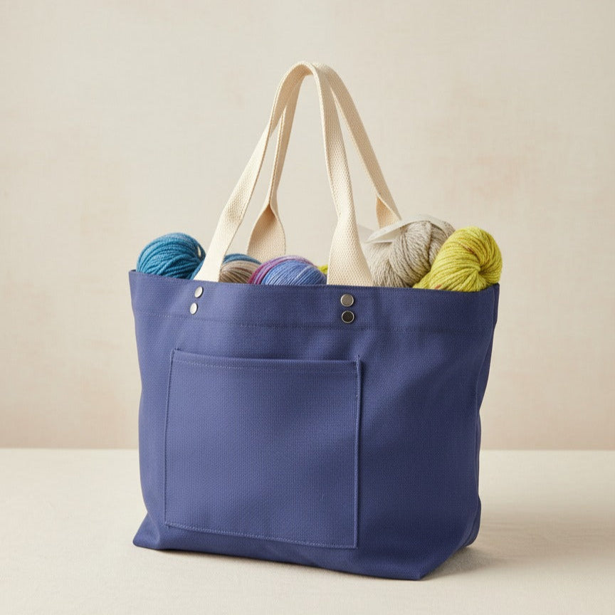 Blue canvas project tote bag for knitters and crocheters.  The bag features an exterior pocket, cream color 2-handle straps, and decorative rivets.  The bag is filled with yarn skeins, on a light neutral color background