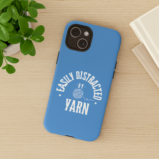 A blue phone case that says "Easily Distracted by Yarn" in white letters on an oak wood table with a green plant and books nearby.  Phone case for knitters and crocheters.