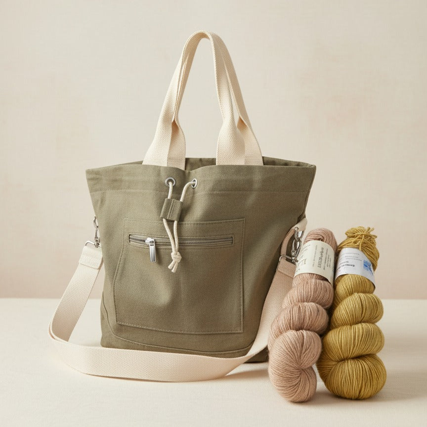 Green canvas project bag with beige accents and two skeins of yarn on a light neutral background.
