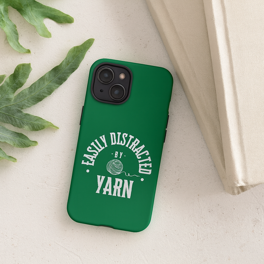 Green phone case with 'Easily Distracted by Yarn' text on a light surface with leaves and a book.