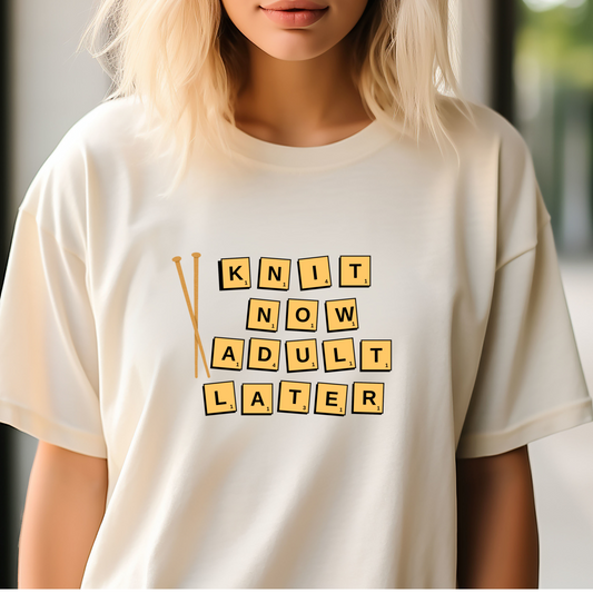 Person wearing a beige t-shirt with a humorous text design that says Knit Now Adult Later in yellow block print.