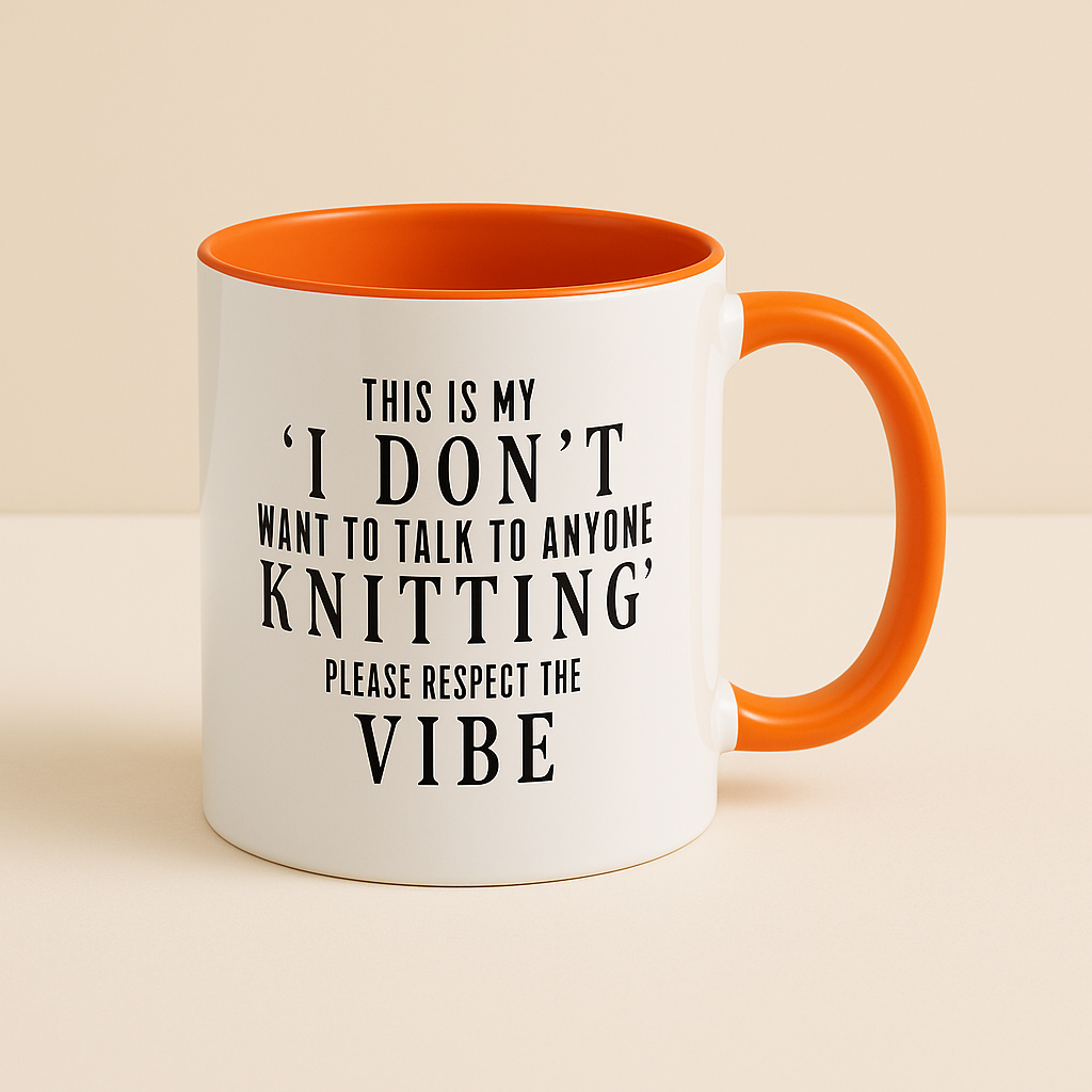 A white ceramic mug with an orange handle and inner lining, featuring bold black text that reads, "This is my 'I don't want to talk to anyone knitting' please respect the vibe." The mug is displayed against a neutral beige background, emphasizing its playful and humorous design. Perfect for knitting enthusiasts who enjoy their quiet time.