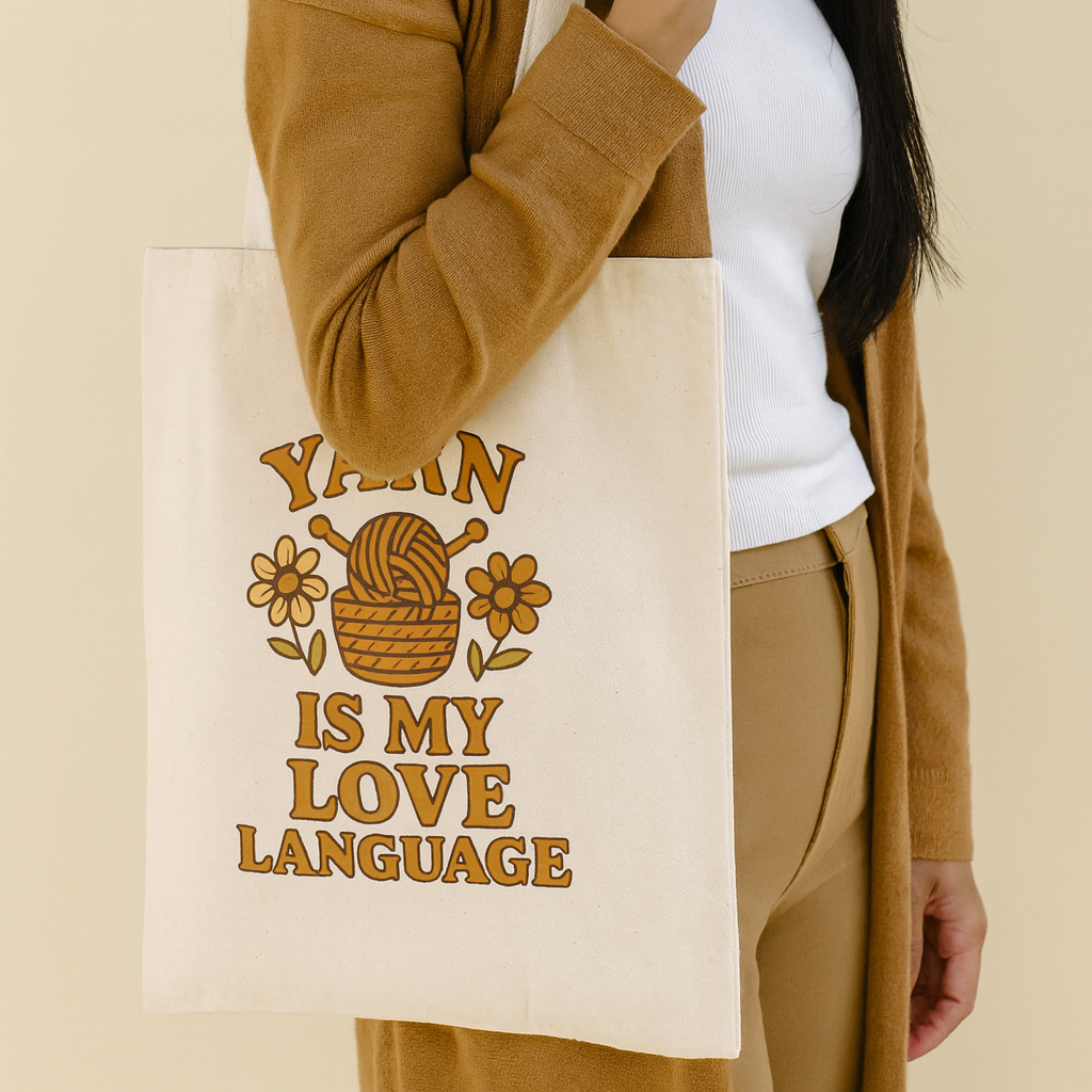 Person holding a natural color canvas tote bag with 'Yarn is my love language' design on the bag.  There a basket with yarn and needles in the design as well as flowers.