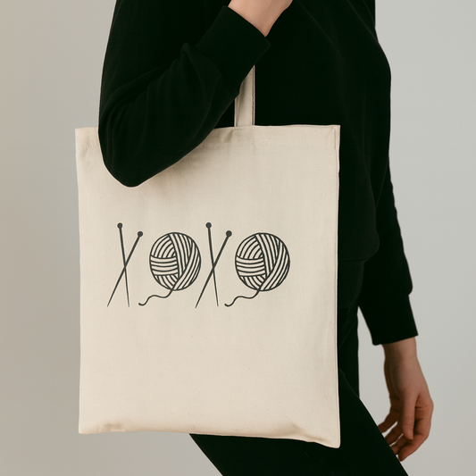 A person wearing black, holding a natural color tote bag with yarn design of knitting needles and yarn on the front.  A play on 'xoxo' text.